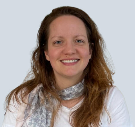 Ashley Miller A smiling woman with long light-brown hair, wearing a white shirt and a patterned scarf, posed against a light blue background.